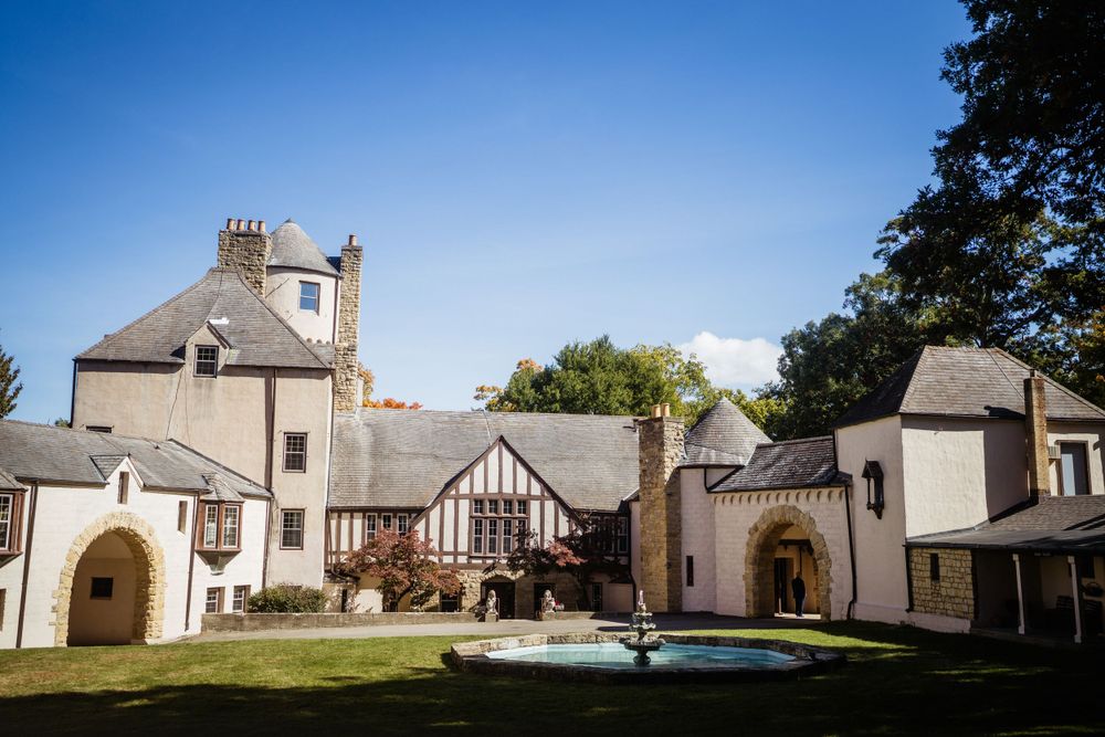Elegant stone manor house with Tudor-style architecture and fountain in courtyard on a sunny day.