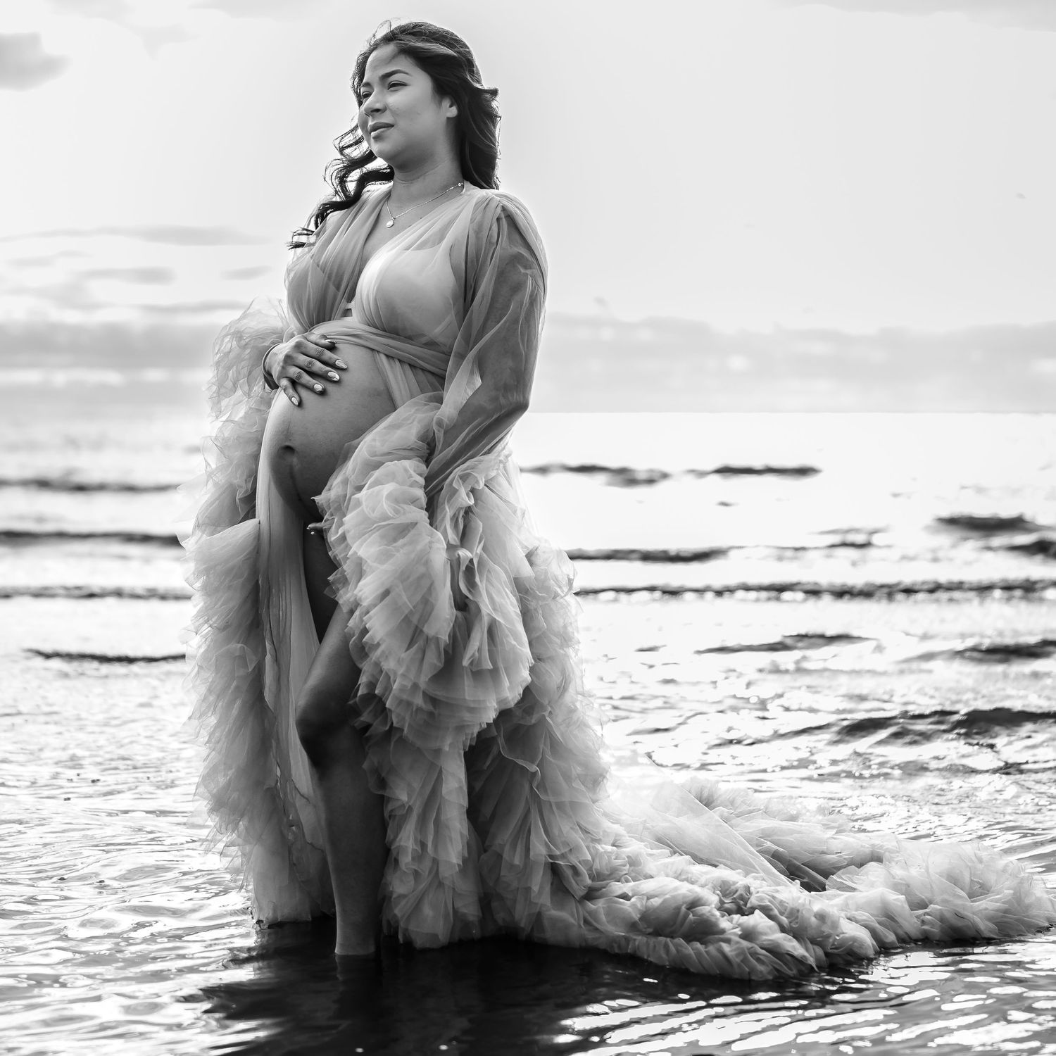 A dramatic maternity photo in black and white showing a pregnant figure in flowing dress at the beach.
