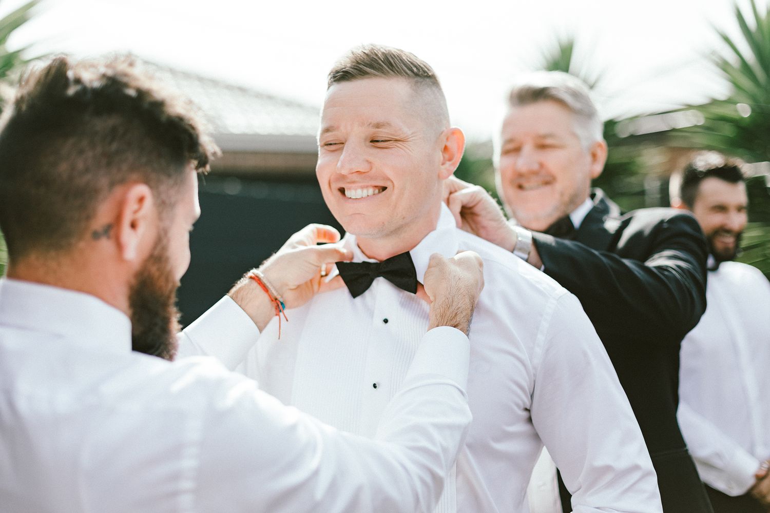 People in formal attire helping adjust a bow tie outdoors.