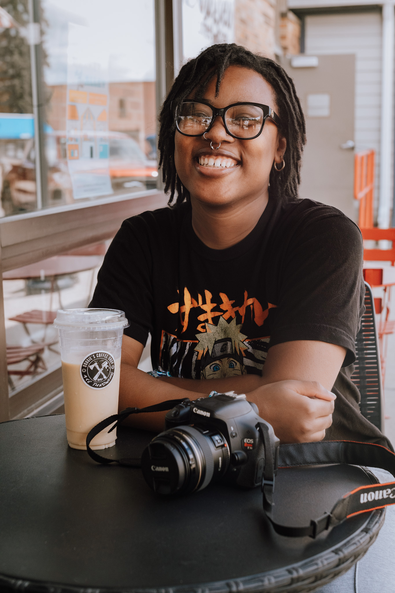 Photographer with glasses relaxes at cafe table with camera equipment and iced drink.