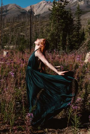Emerald dress twirls among purple wildflowers with mountain peaks in background.