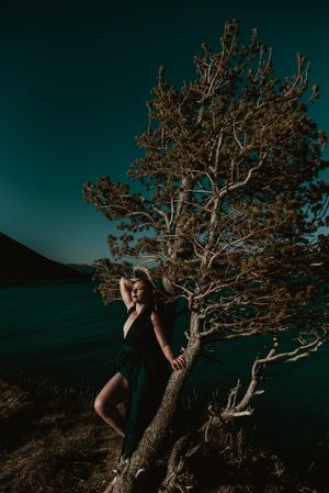 Moody portrait of a woman against gnarled tree with dark lake and mountains in background.