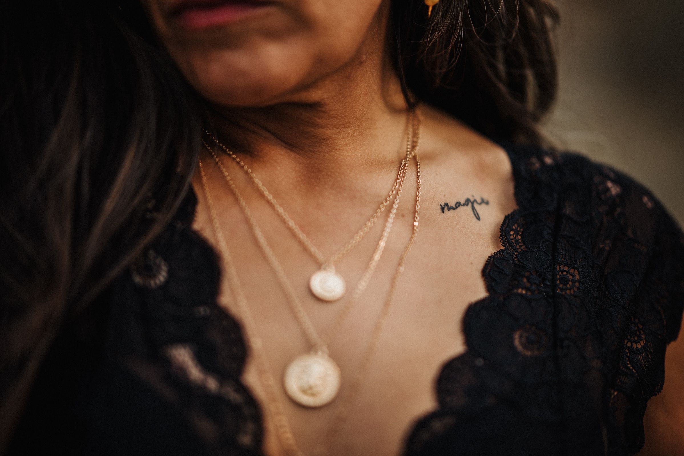 Close-up of layered necklaces and lace detail on black clothing with a tattoo that says magic.