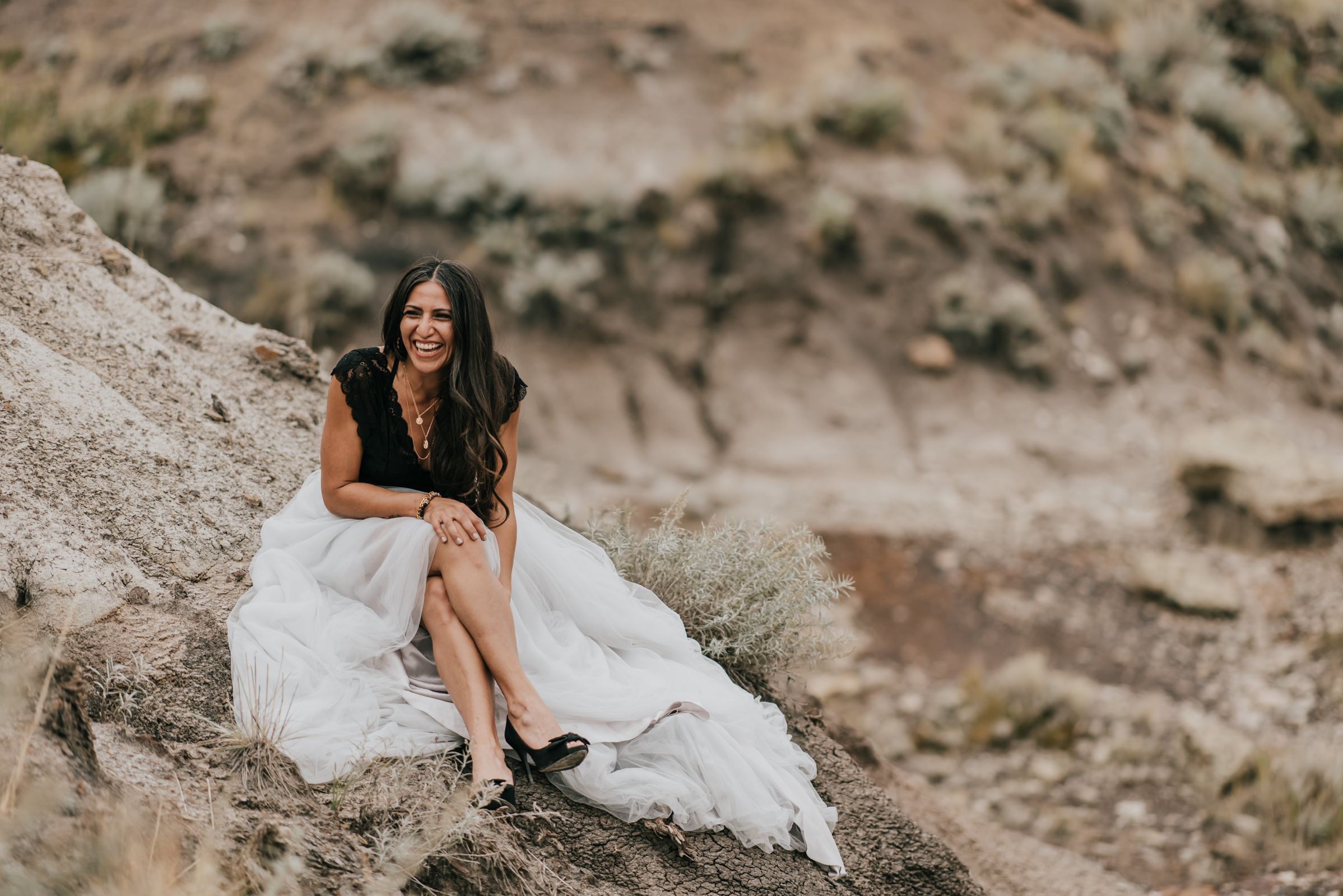 Person in black top and white dress sits elegantly on rocky cliff during photo shoot.