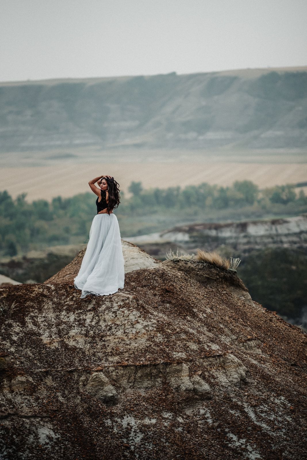 Figure in flowing white dress stands atop rocky hill against misty mountain landscape at dusk.