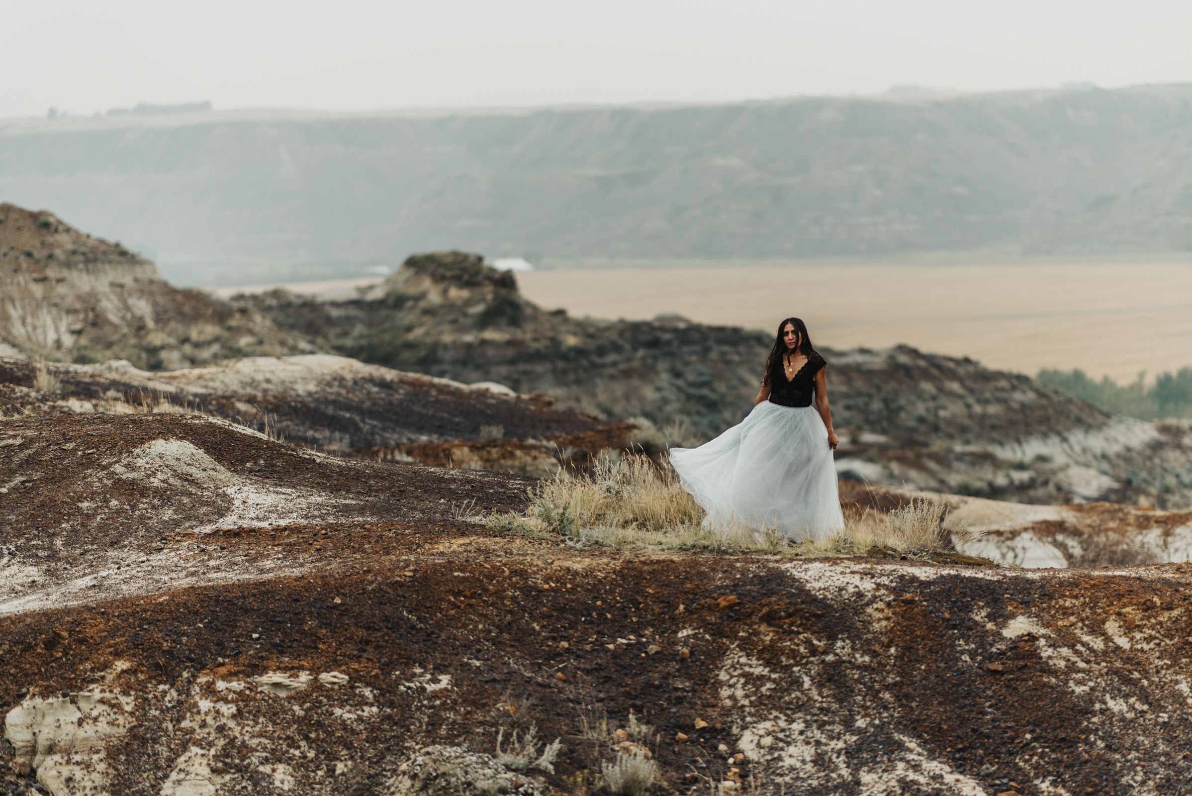 Figure in flowing white dress stands on rocky overlook with mountain vista in background.
