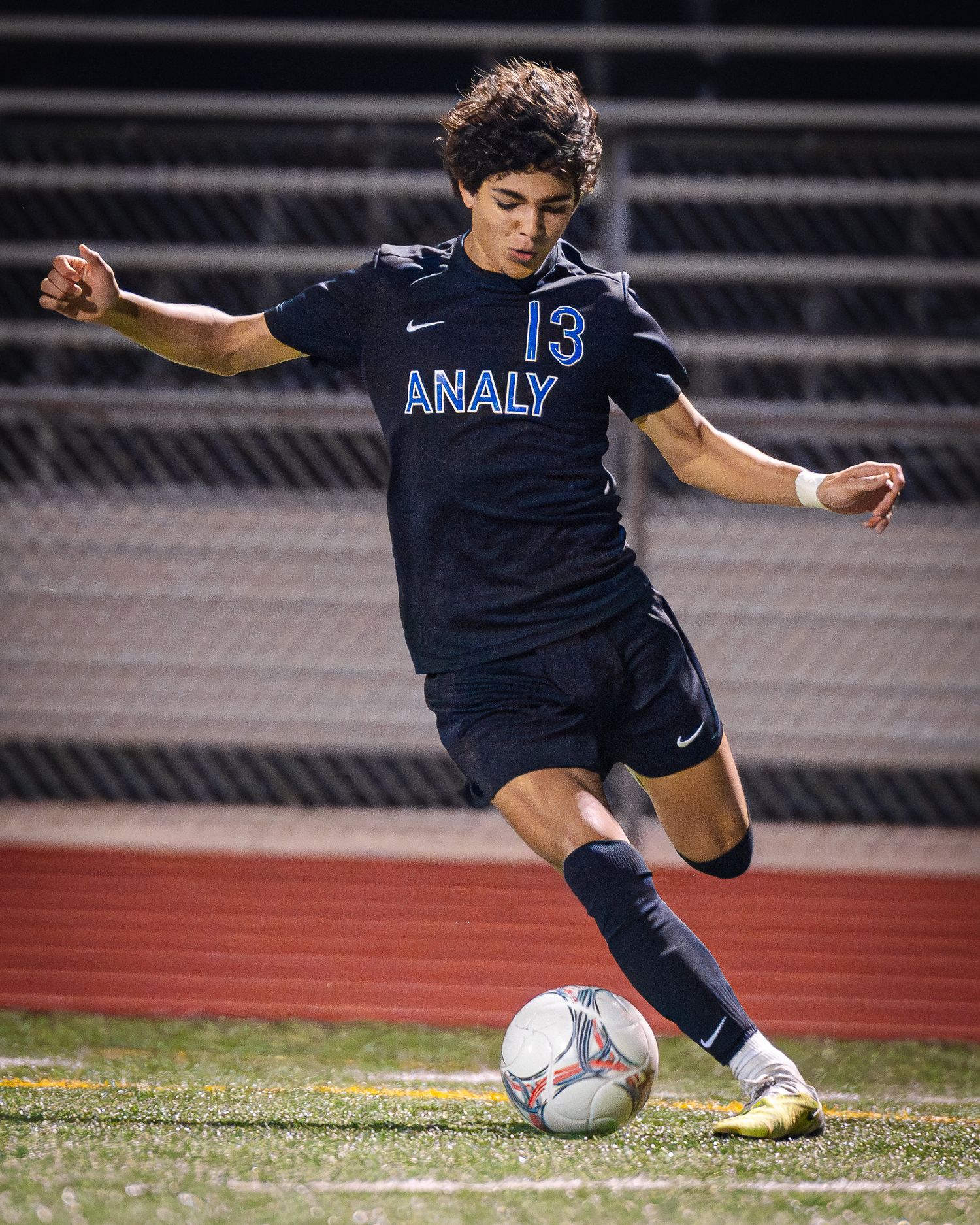 Soccer player in navy Analy jersey kicks ball during match on red turf field.