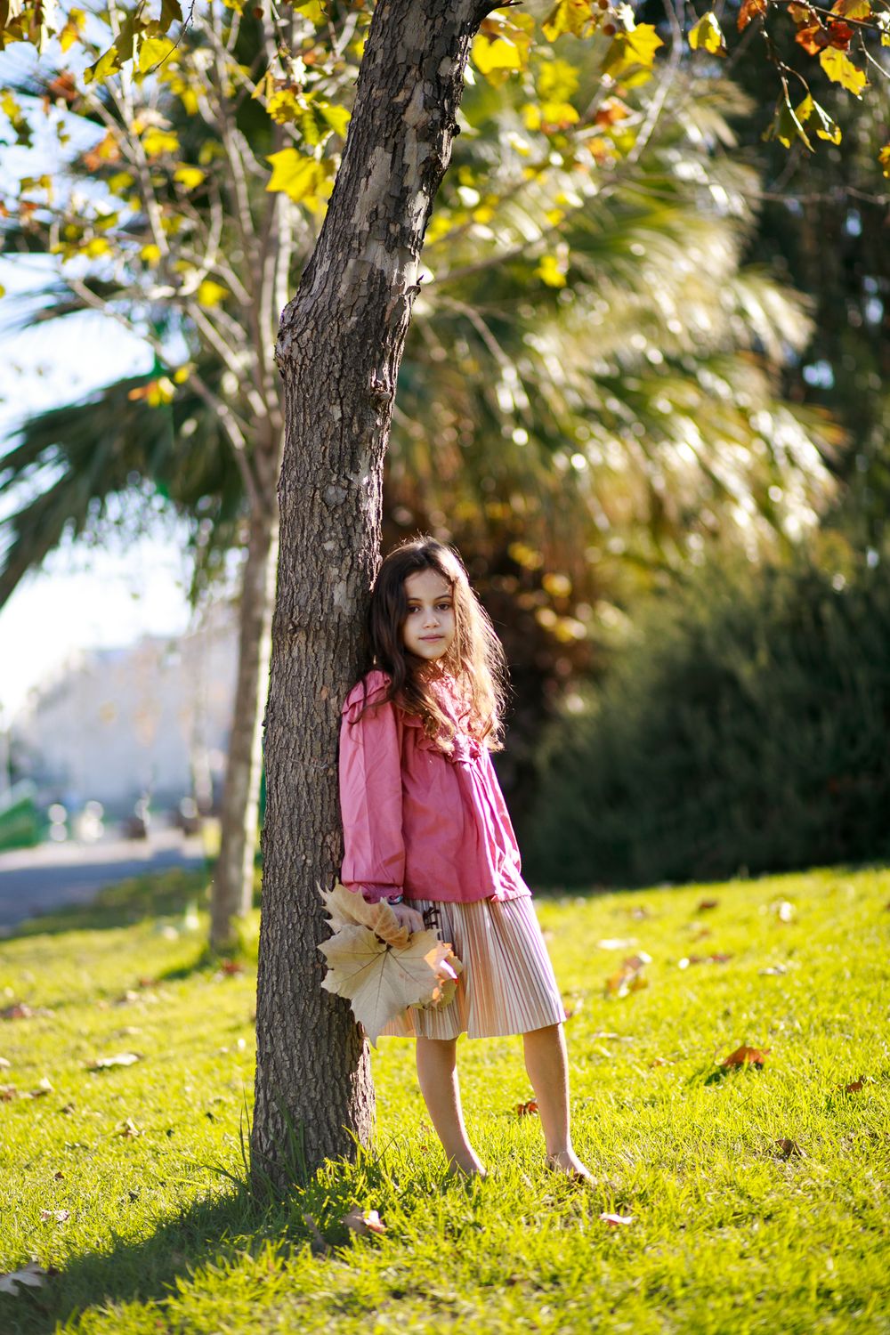 A picture of a beautiful girl leaning against a tree in the garden with a bouquet of fallen leaves in hand