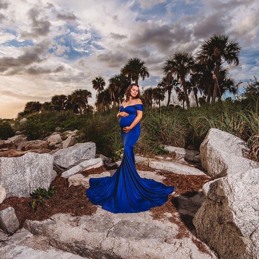 Maternity photo in royal blue dress among palm trees and rocks at sunset.