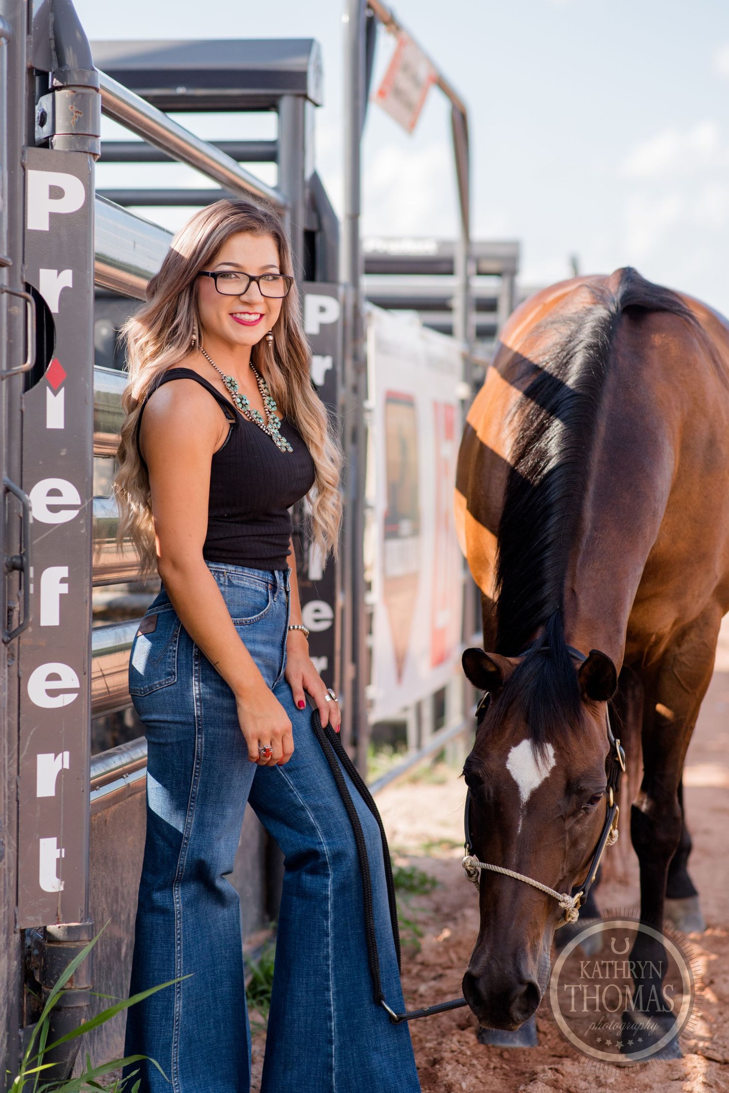 Someone in retro-style denim and glasses poses with a grazing horse by metal stalls.