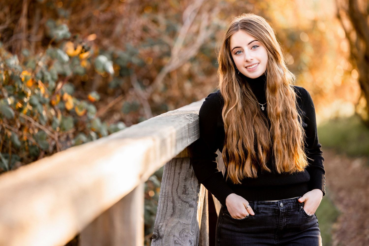 Teen girl with long auburn hair smiling by wooden fence in fall.