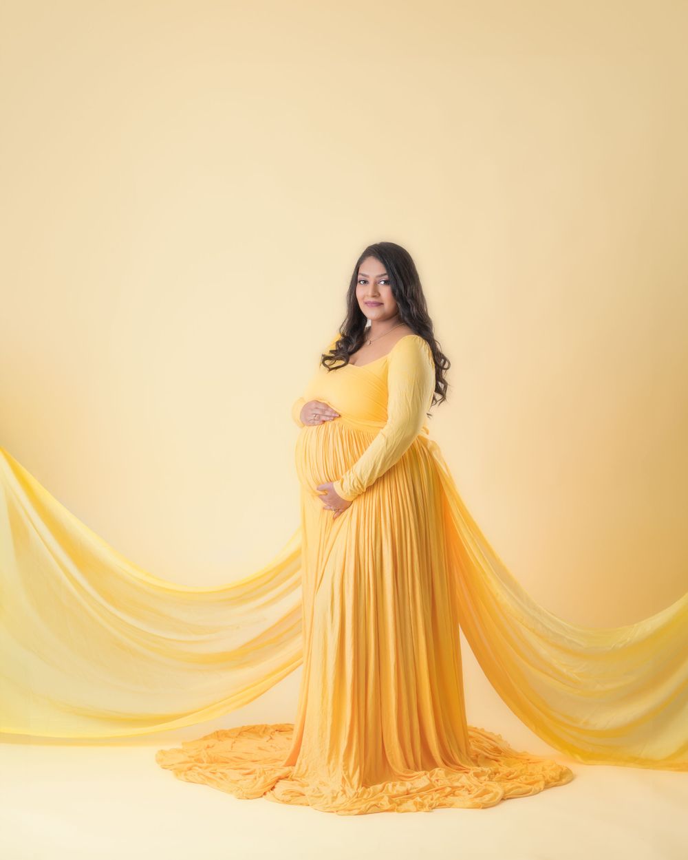 Maternity photo of a person in a flowing yellow gown against a warm beige backdrop.