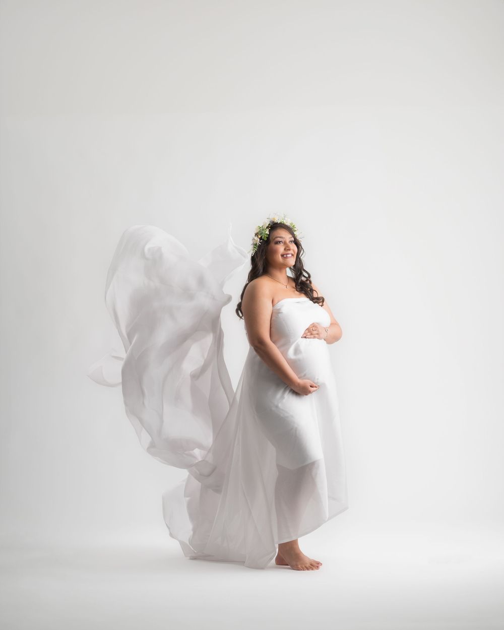 Maternity photo of expectant mother in flowing white dress against light background.