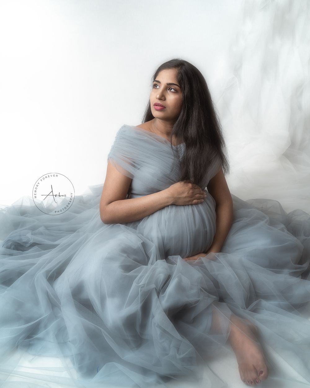 An ethereal maternity portrait featuring a flowing gray tulle gown against a white background.