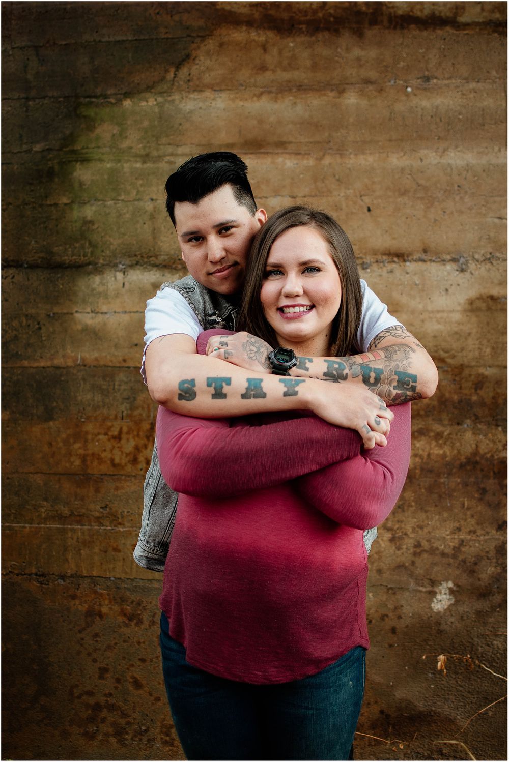 Tattooed couple sharing warm hug and smiles against rustic concrete wall.