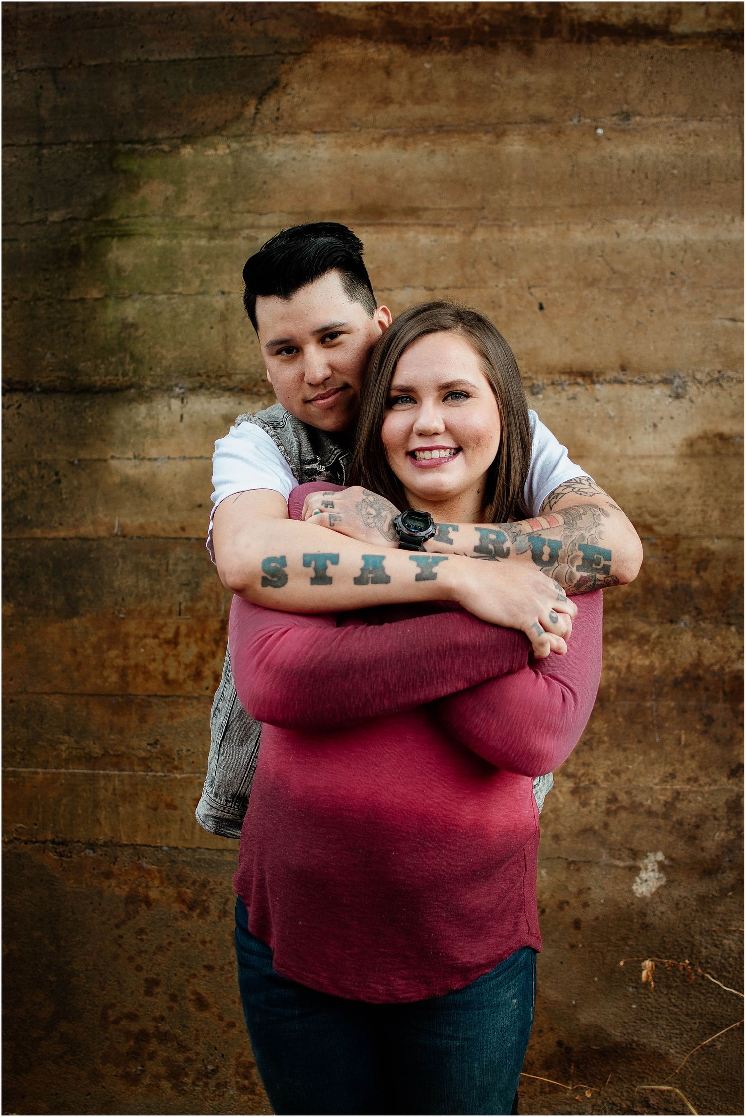 Tattooed couple sharing warm hug and smiles against rustic concrete wall.