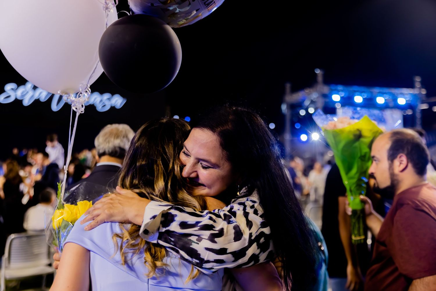Two women share emotional hug at outdoor celebration with balloons and sunflowers.