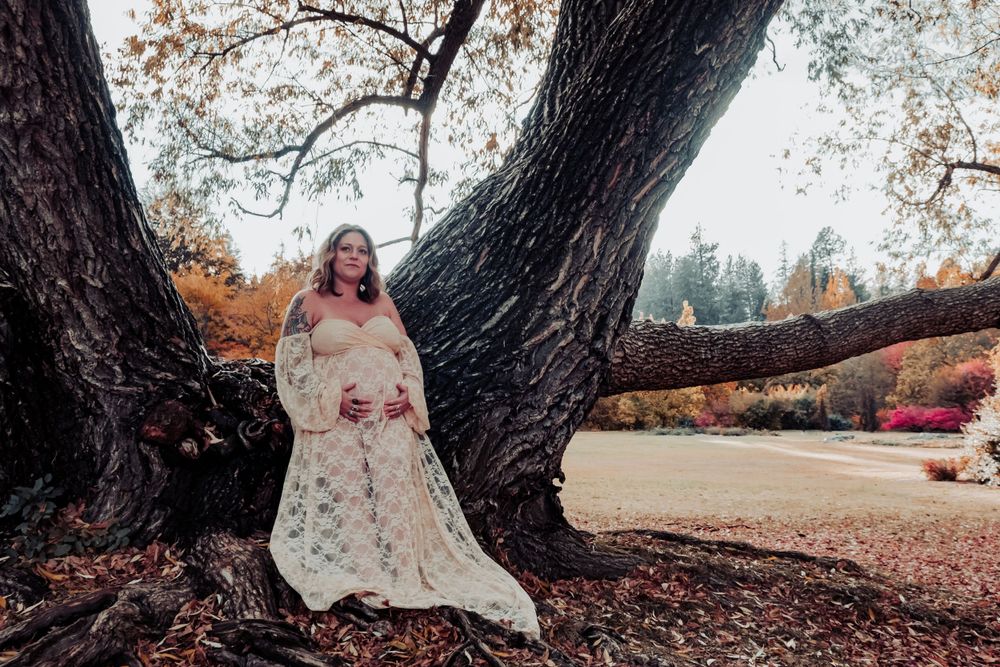A bride in an ornate lace wedding dress poses against a large tree trunk in an outdoor garden setting.