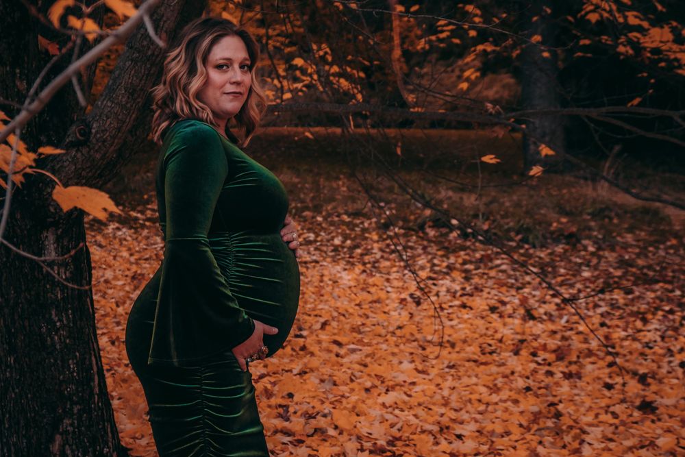 Autumn maternity portrait in green velvet dress surrounded by fallen leaves.