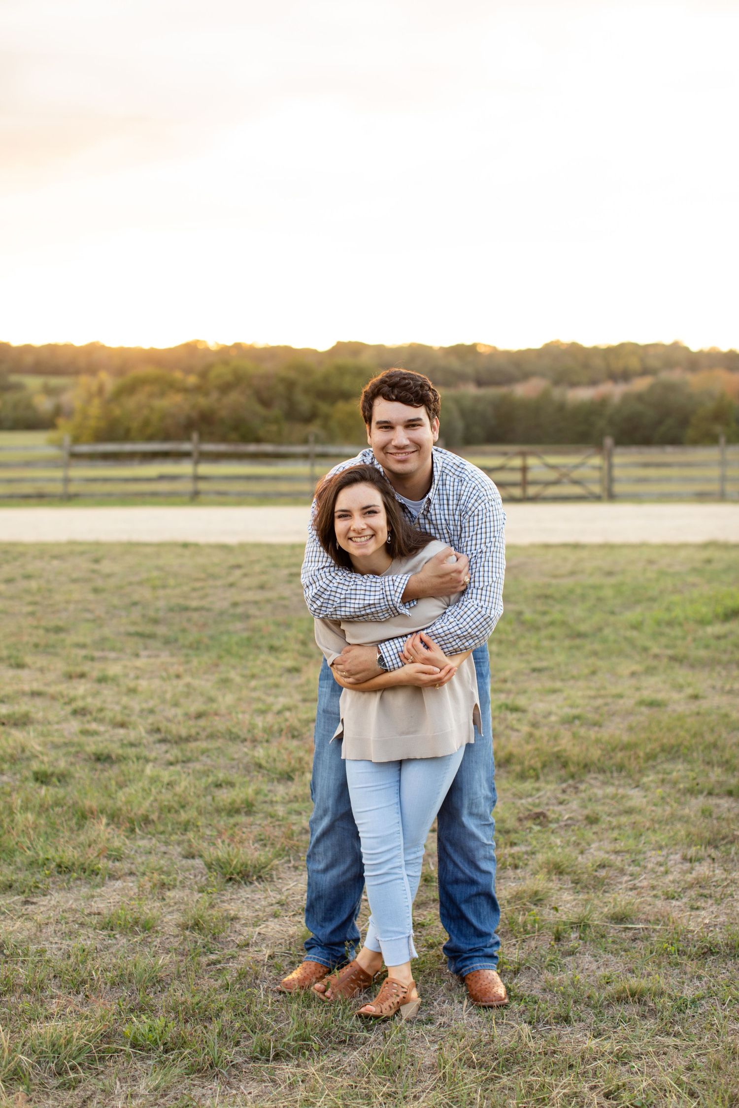 Couple sharing a warm embrace while standing in a grassy field with fence and sunset in background.
