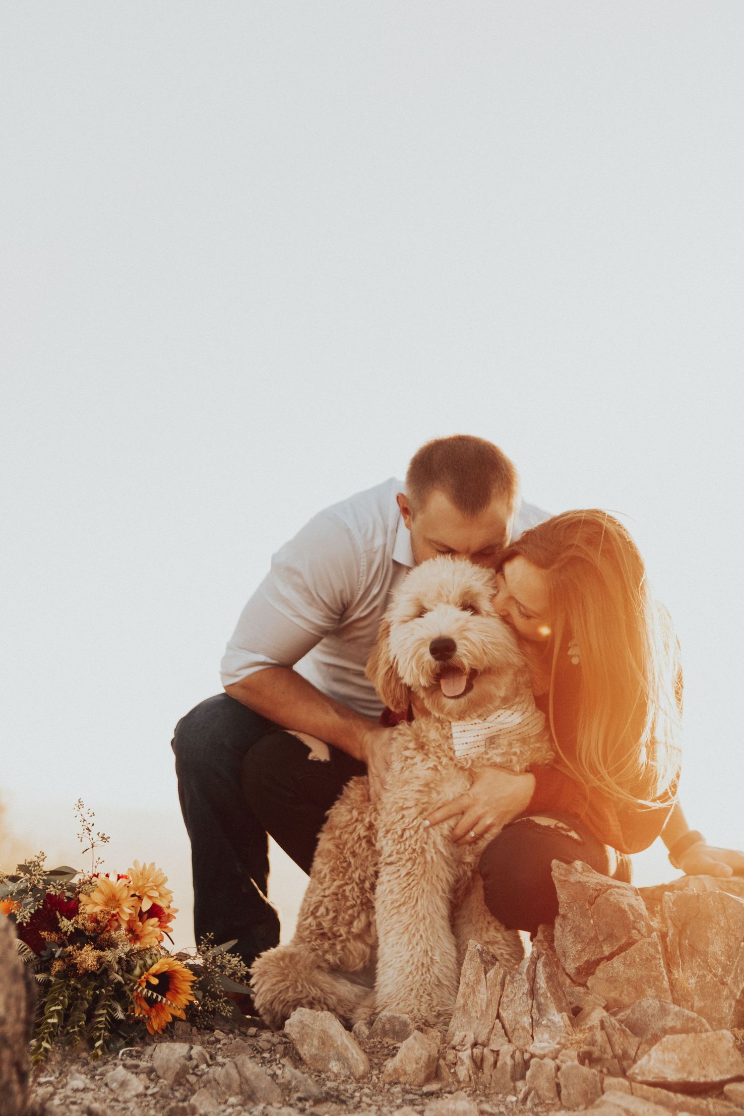 happy couple snuggling their goldendoodle as the sun rises in the background. beautiful flowers peek out of the left