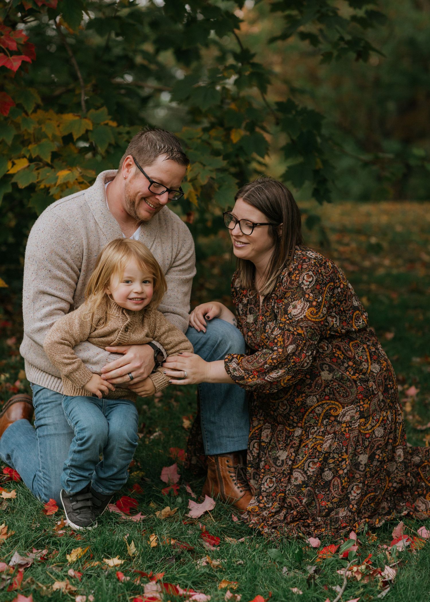 A family sits together on grass surrounded by fall foliage as red leaves scatter around them.