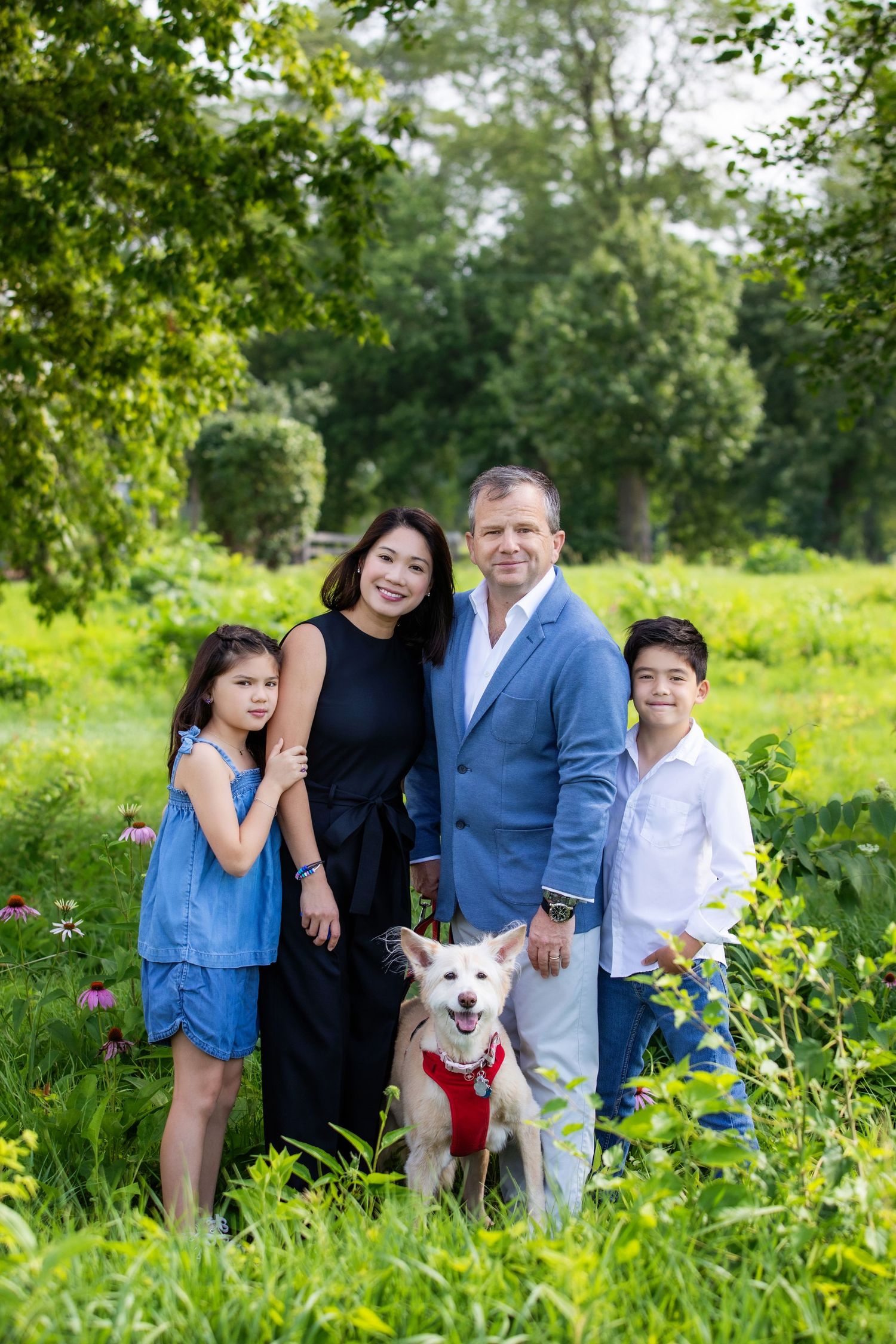 Family of four with white dog posing together in sunny outdoor garden setting.