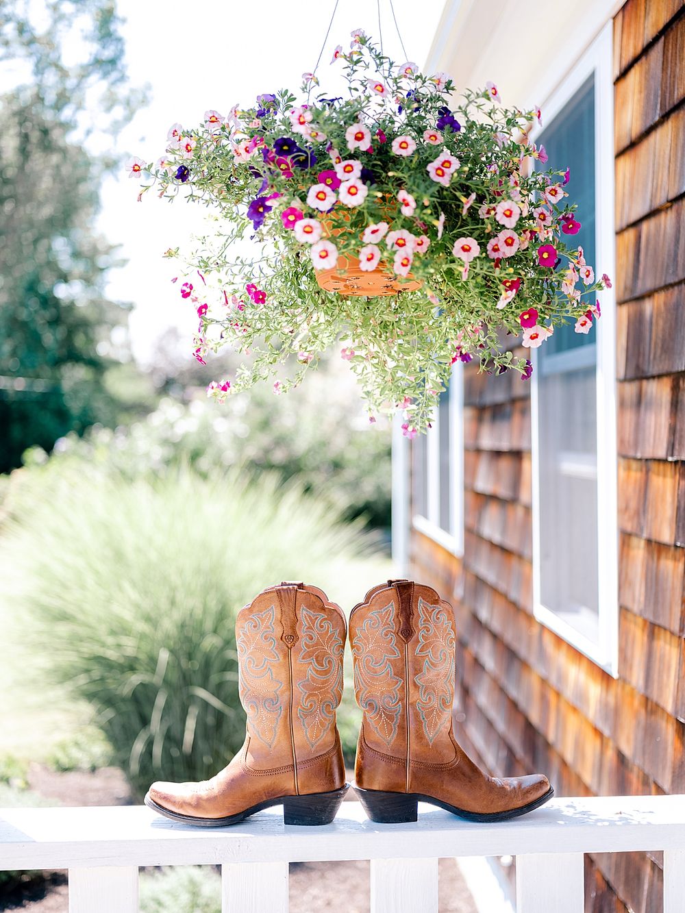 Brown leather cowboy boots beneath a hanging basket of pink and purple flowers on a porch.