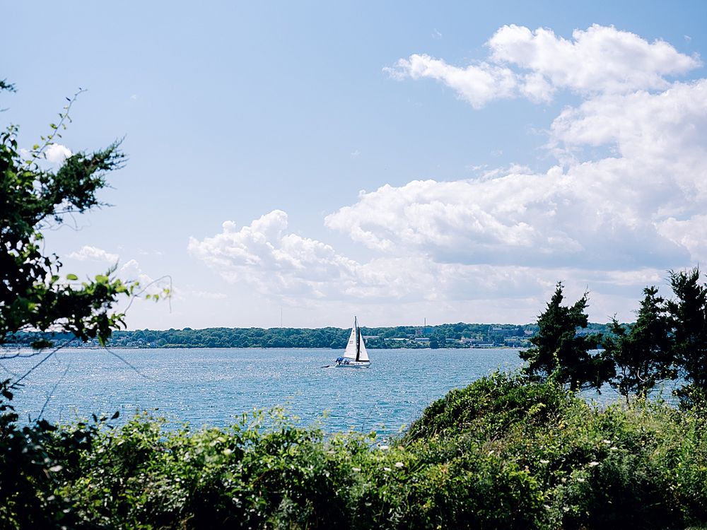 Sailboat on blue water with evergreen trees and white puffy clouds framing the coastal view.