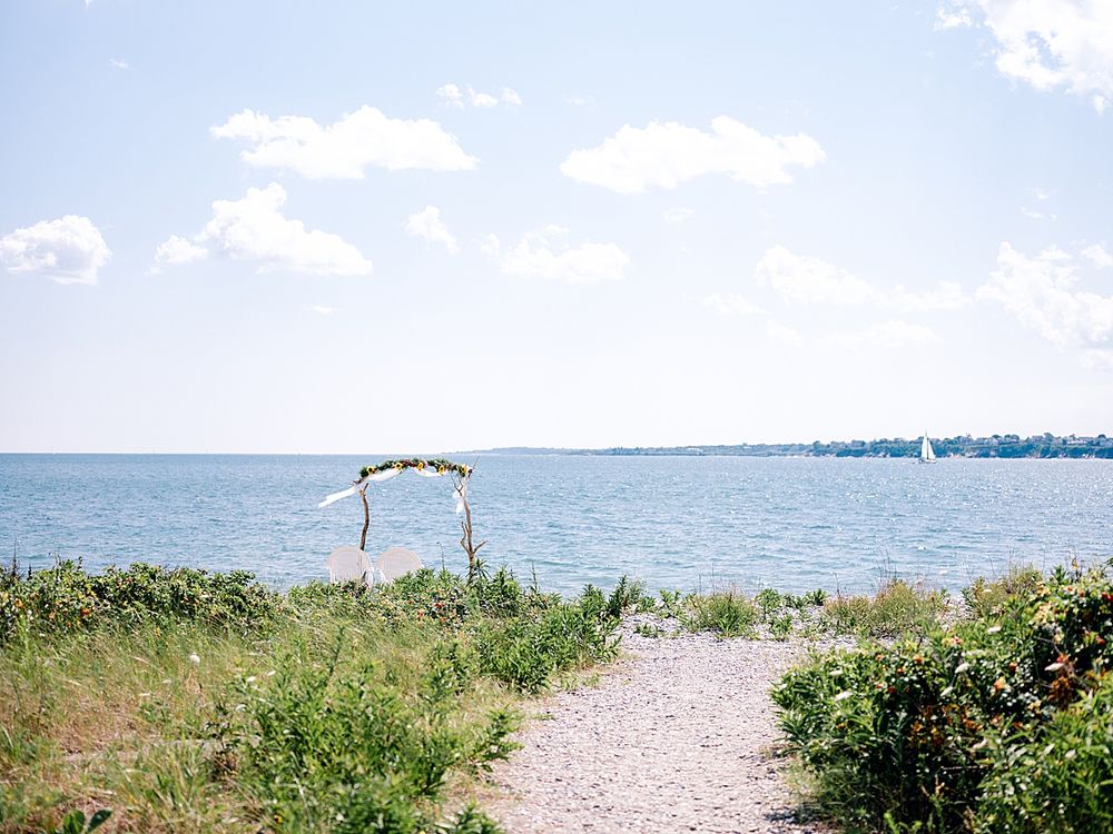 Simple driftwood wedding arch on beach path with ocean view and blue skies.