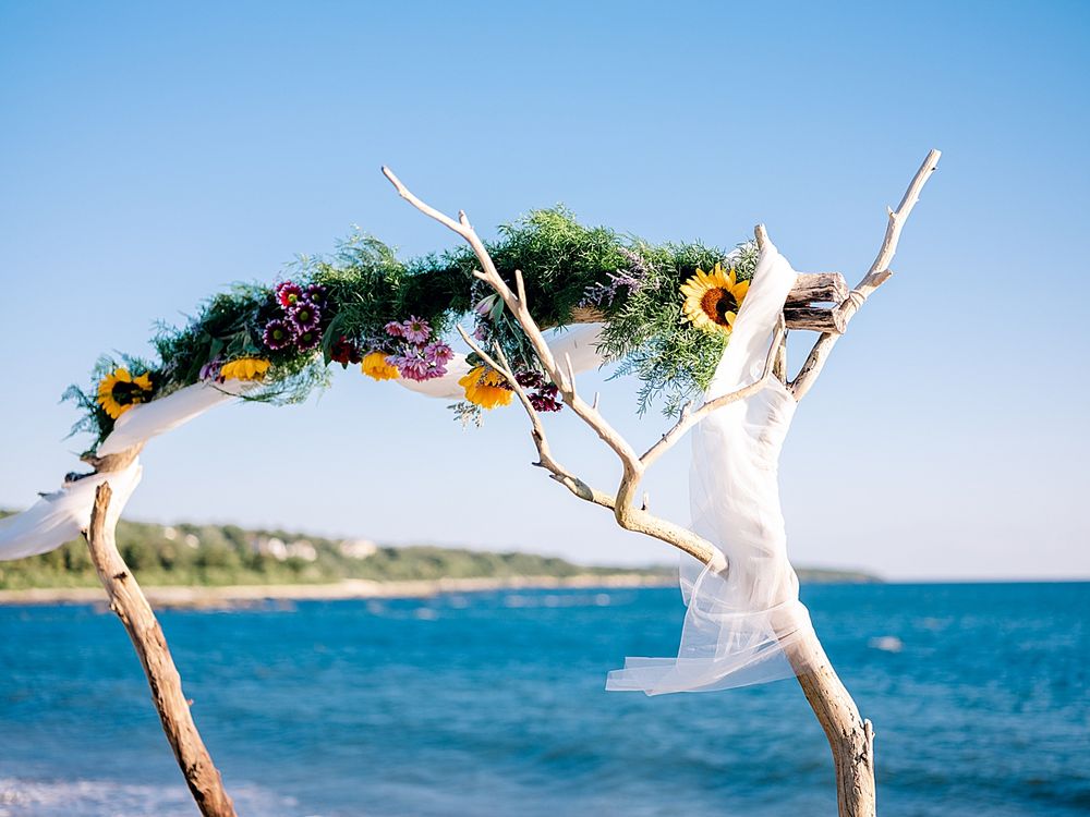 Floral decorated driftwood arch against blue ocean backdrop with white fabric flowing.