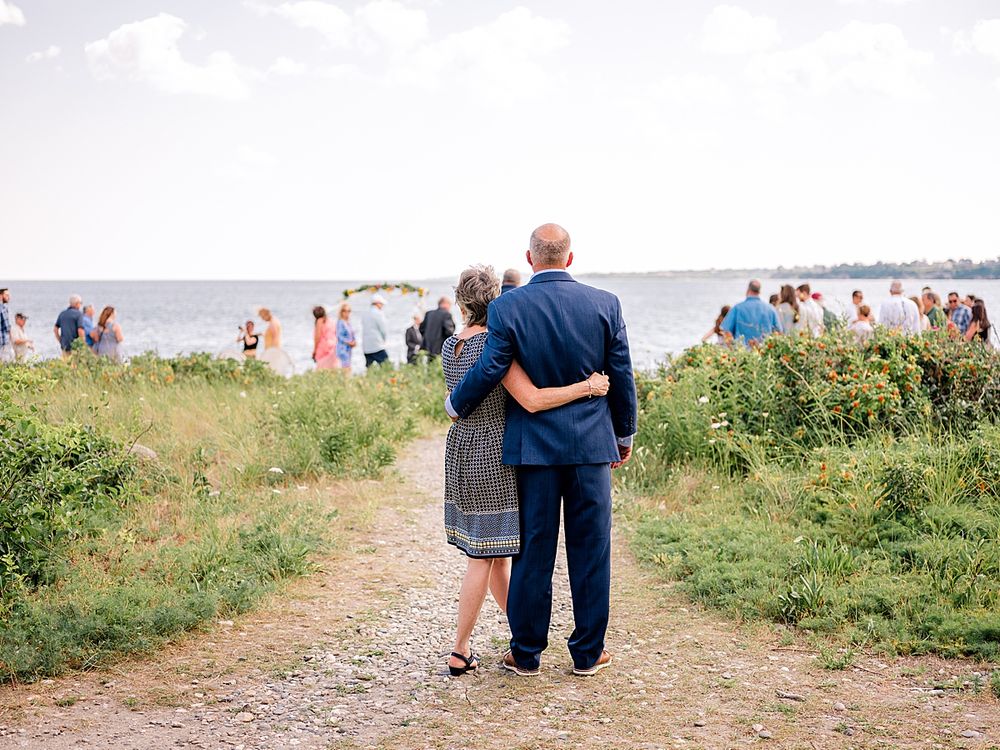 Couple walking down coastal path with ocean and wedding guests visible in background.