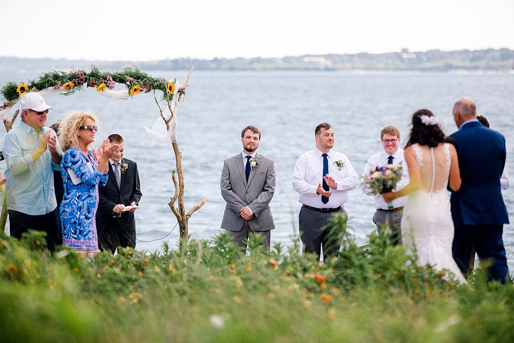 Outdoor beach wedding ceremony under floral arch with ocean backdrop.