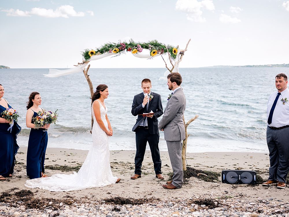 An outdoor beach wedding ceremony with attendants in navy blue dresses gathered under a driftwood arch with flowers.