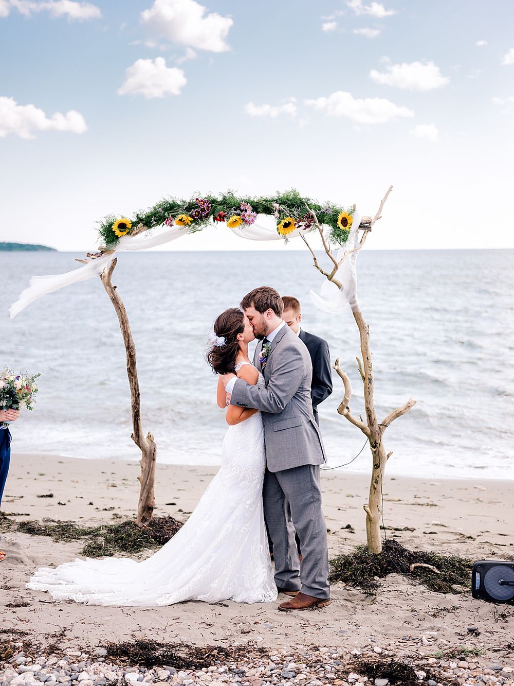 Newlyweds embrace under a rustic floral arch on a beach with ocean waves in the background.