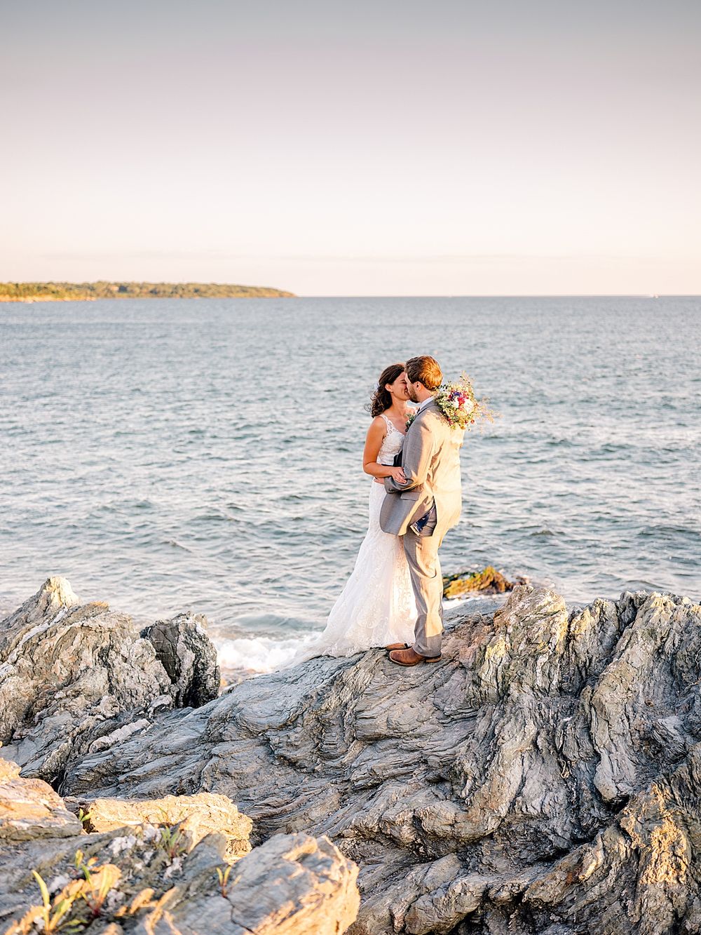 Couple in wedding attire embracing on rocky oceanside cliffs during golden hour.