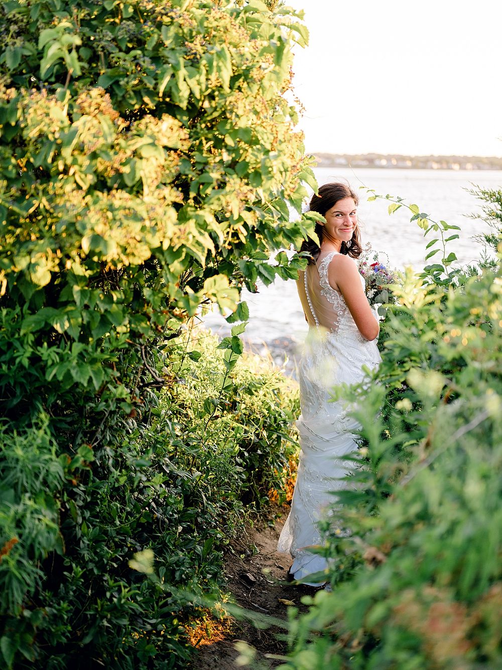 Elegant bride in lace gown poses among lush green foliage near waterfront.