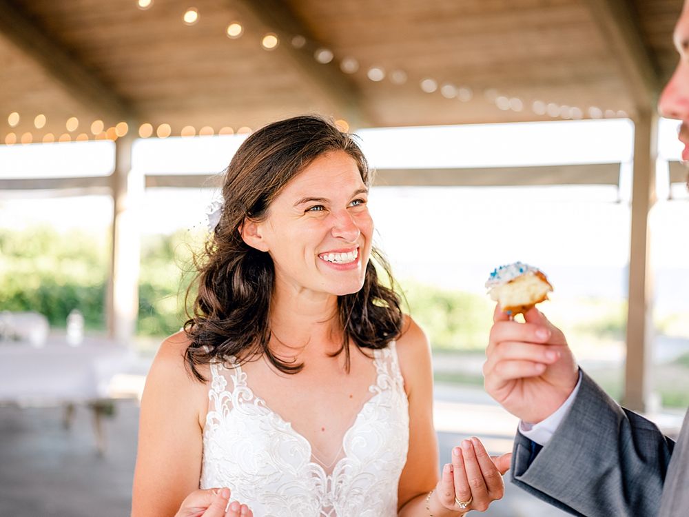 A bride in a lace wedding dress laughs joyfully while holding a cupcake under string lights.