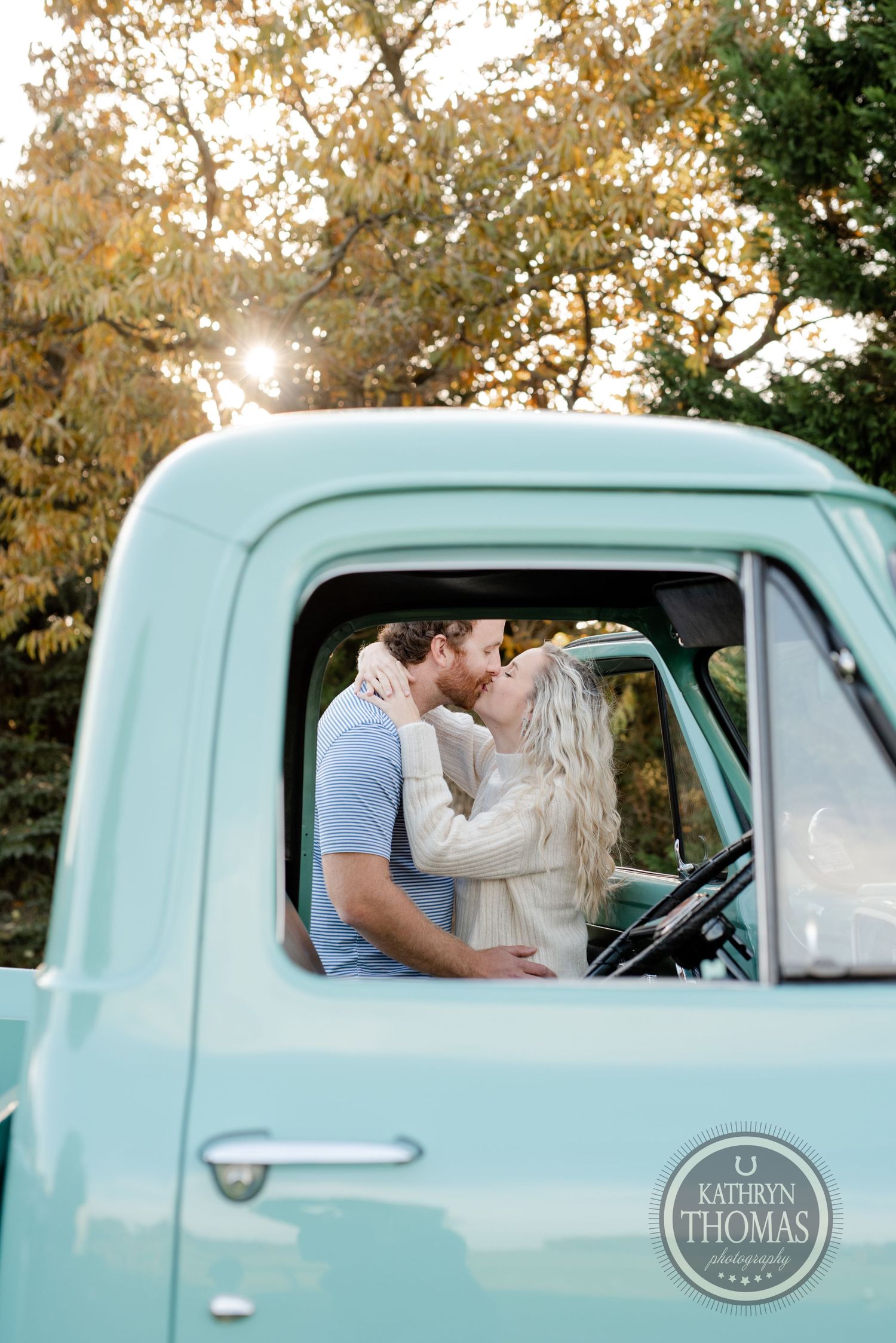 Intimate moment captured between couple in vintage turquoise truck with sunlight peeking through trees.