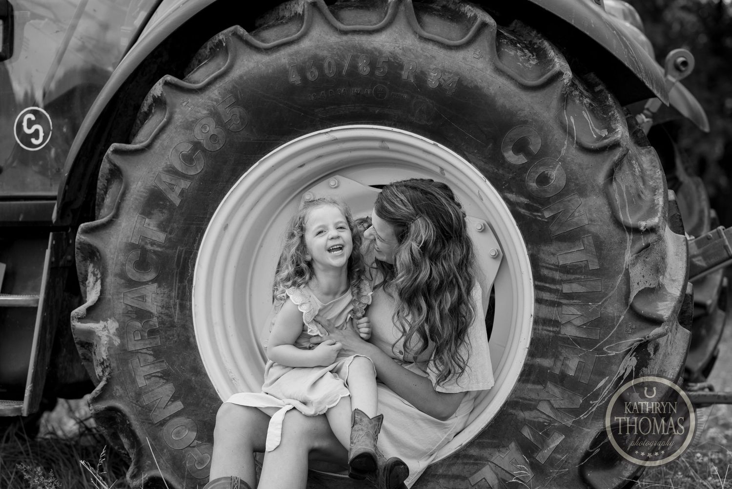 A black and white portrait of two people sitting inside a large tractor tire wheel.