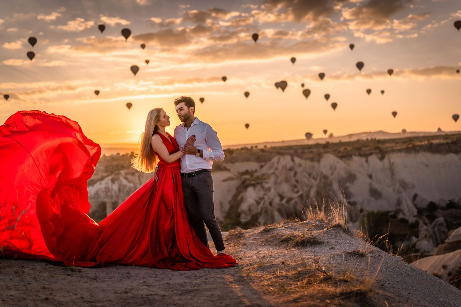 Red flowing dress catches wind as couple poses against backdrop of floating balloons at sunset.