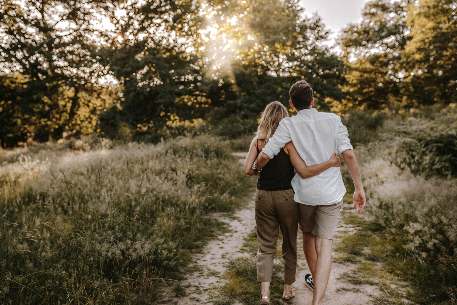 A couple walks arm in arm down a sunlit path through golden grass at sunset.
