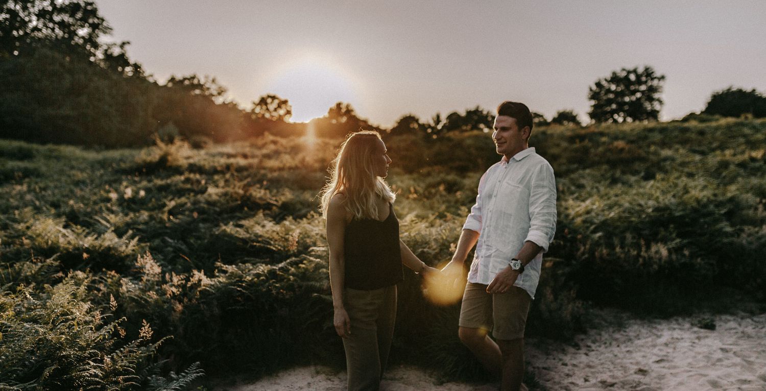 A couple stands together in a meadow during golden hour with sunlight filtering through trees.