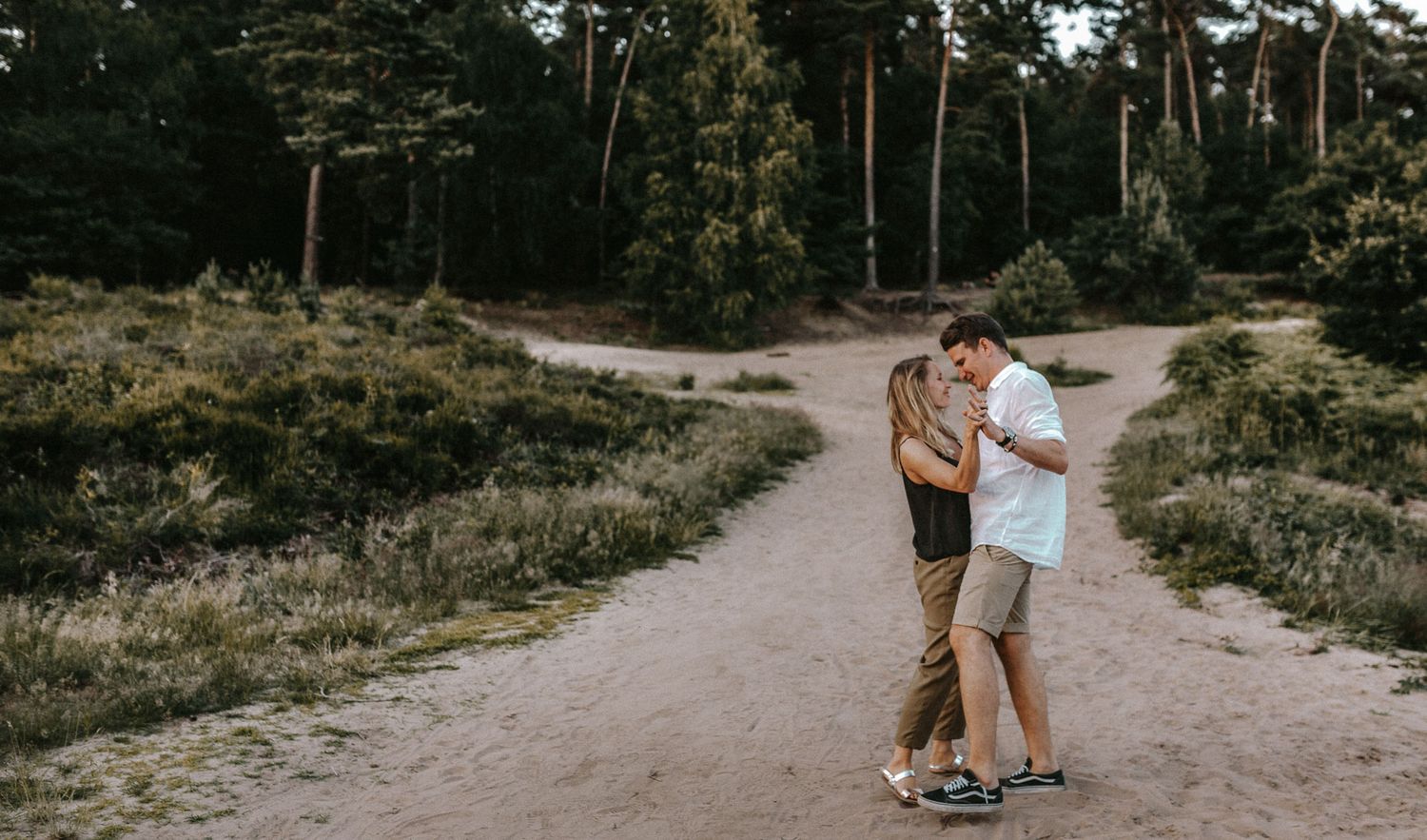 A couple embraces on a winding forest path surrounded by pine trees.