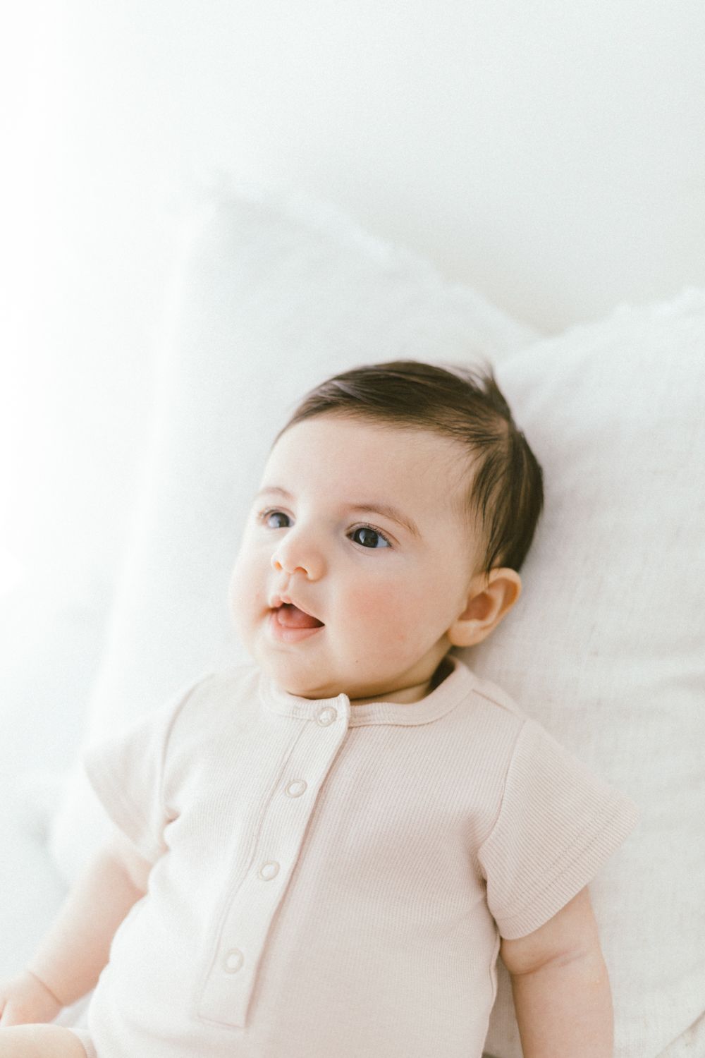 Infant in light pink outfit gazes upward while resting on white linens.