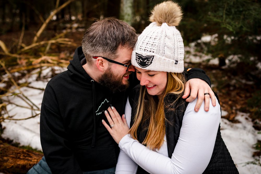 Winter engagement photos featuring cozy moments and white knit hat details in snowy forest setting.
