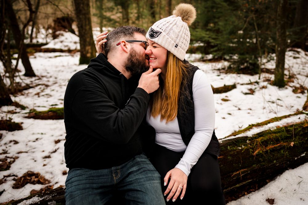 Candid winter engagement photos showing playful and romantic moments in snow-covered woods.