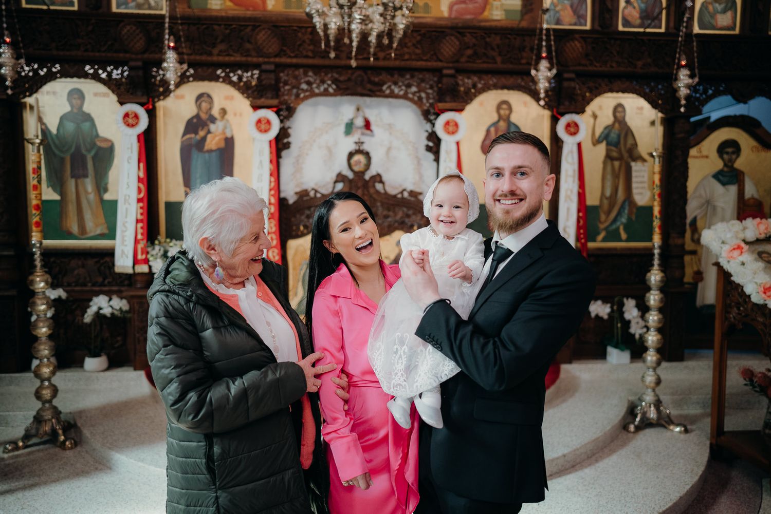 grandma, mum and dad smiles as baby in christening gown smiles at the camera