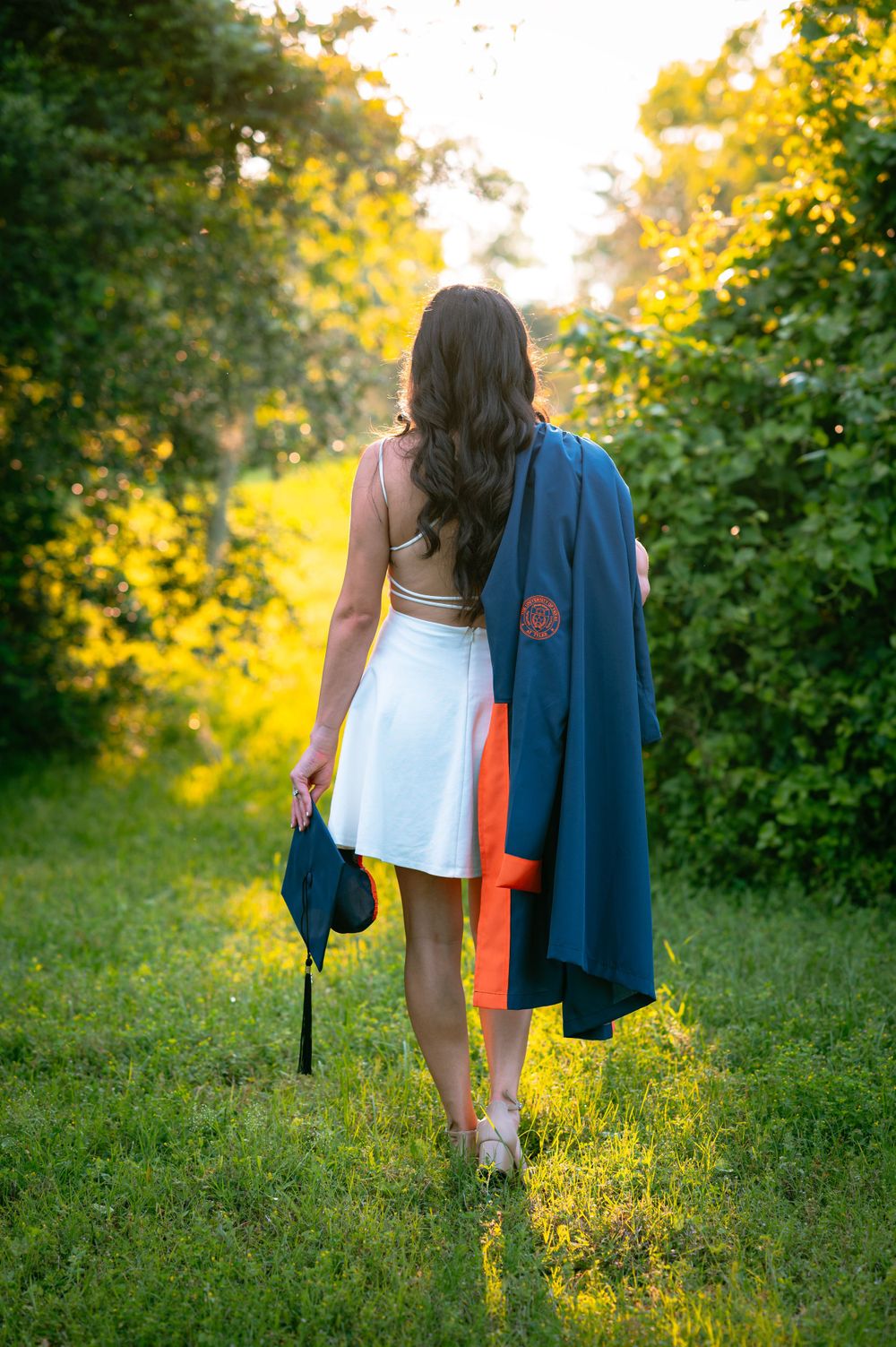 Graduate walking through grass at sunset carrying academic regalia and diploma.