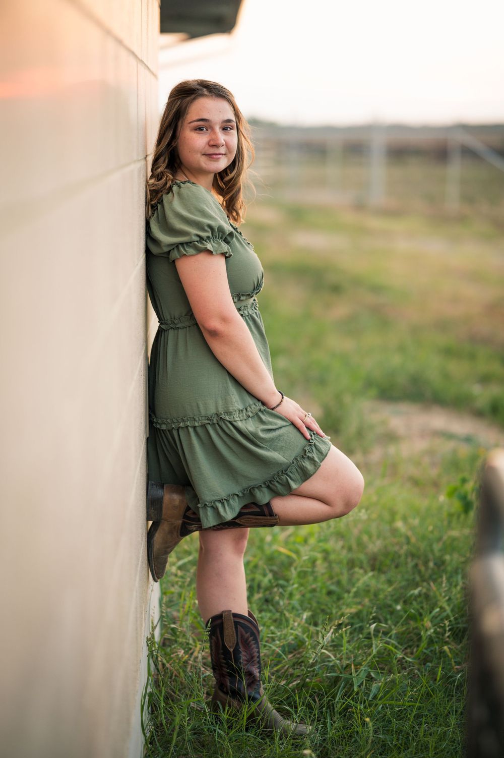 Someone in a green dress and brown boots poses against a white wall in a grassy field.